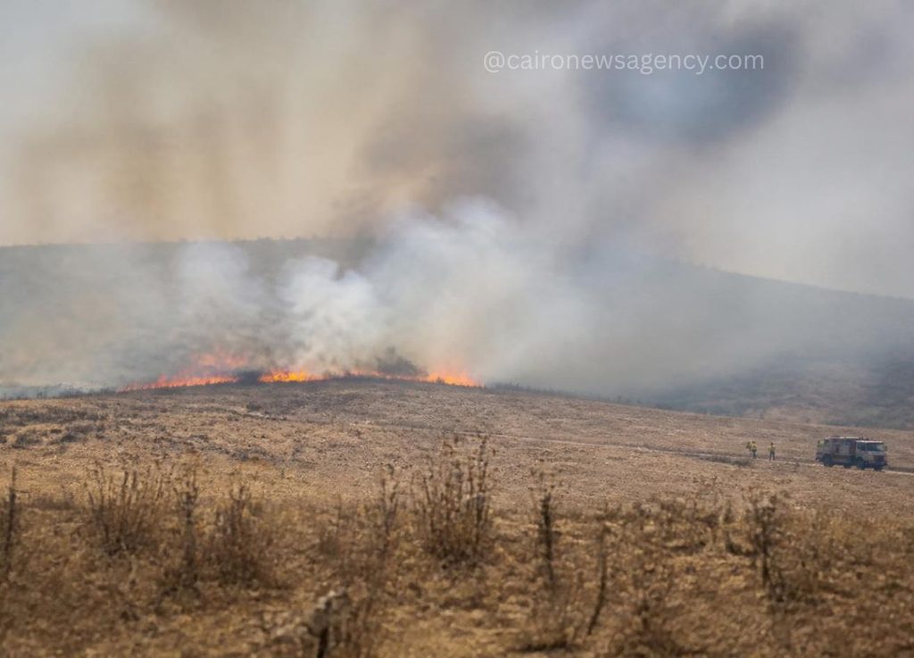 Firefighters and a fire truck in the distance respond to a small wildfire burning along a dry hillside with orange flames and rising smoke.
