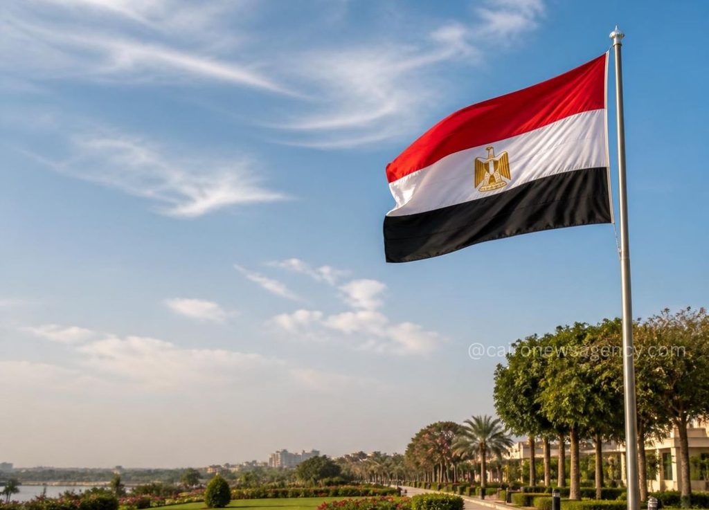 Egyptian flag waving on a tall flagpole beside a palm-lined park under a blue sky with a distant waterfront.