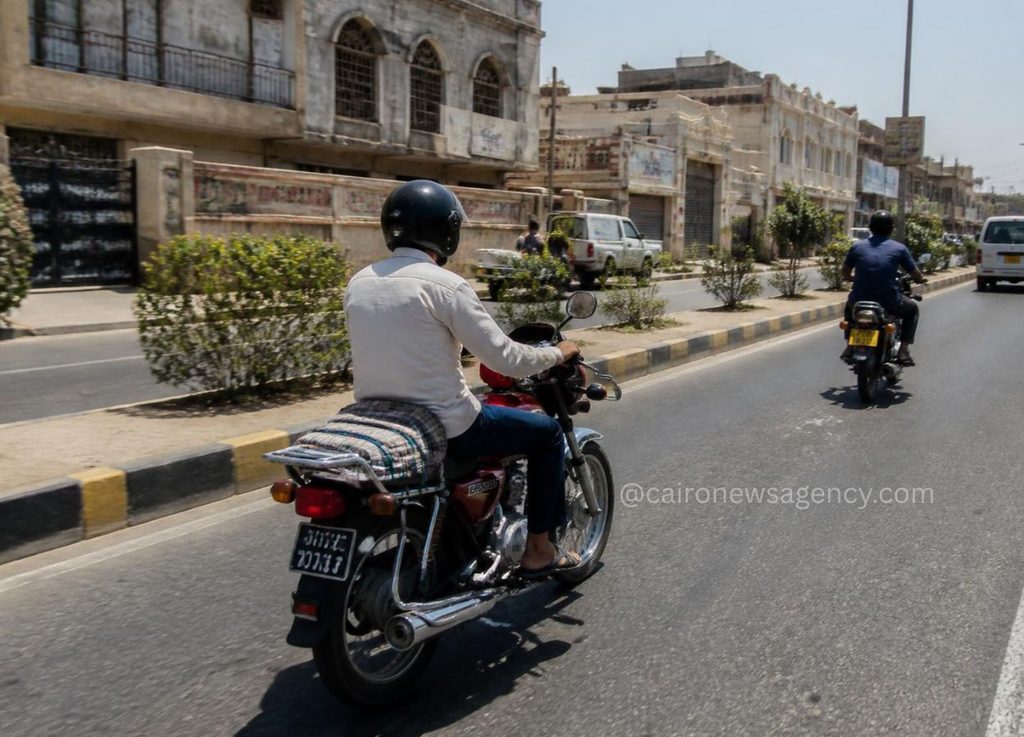 Two men ride motorcycles on a city street; foreground rider wears a white sweater and helmet with a rolled mat strapped to the back near a yellow–black curb.