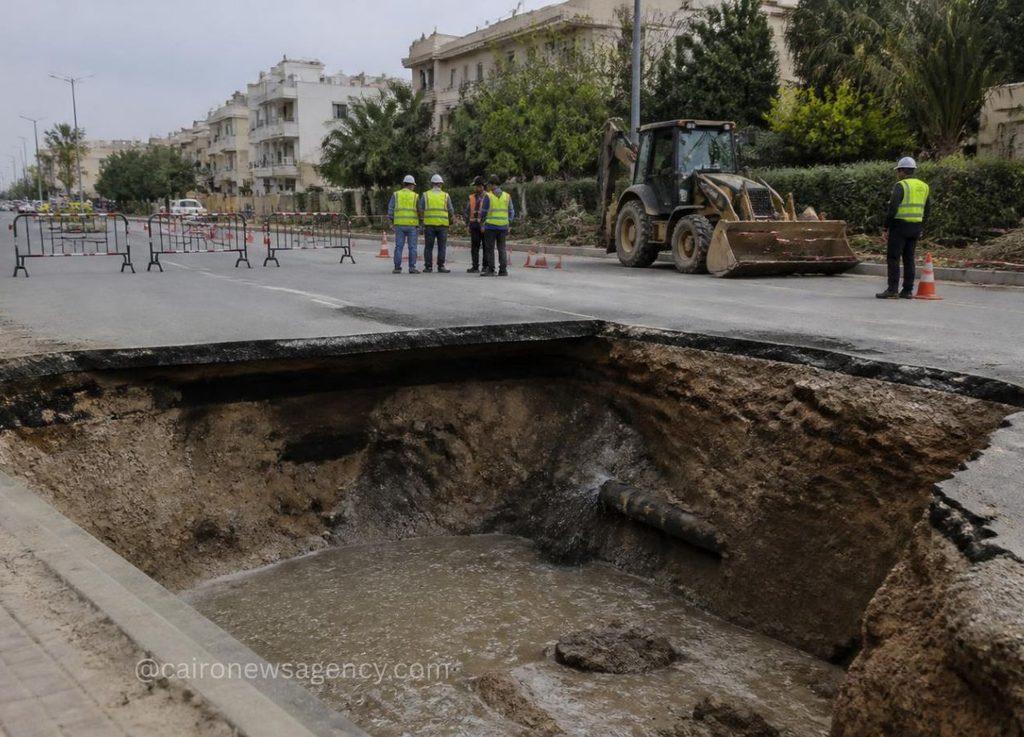 Sinkhole opens in a city street as construction workers in high-visibility vests assess the area with a backhoe nearby and barriers around the site.