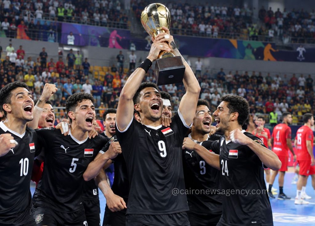 Egyptian men's volleyball team celebrates, lifting a trophy in a stadium with cheering crowd behind them.